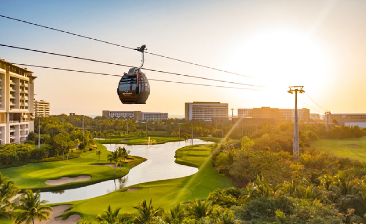 Un teleférico sobre un campo de golf con fuente de agua, rodeado de vegetación y edificios.