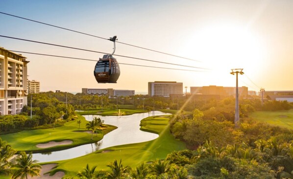 A cable car over a golf course with a water feature, surrounded by greenery and buildings.