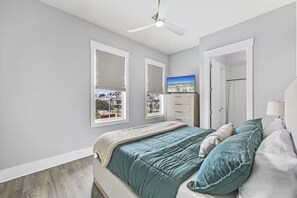 Ein Schlafzimmer mit einem Bett, einem Deckenventilator, zwei Fenstern mit Jalousien, einem Fernseher und einer Tür zu einem weiteren Raum.