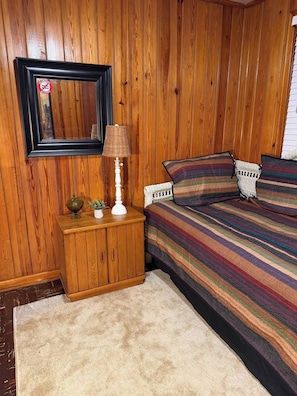 Een slaapkamer met een houten wand, een bed met gestreepte beddengoed, een nachtkastje met een lamp en een spiegel.