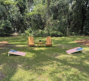 Two Adirondack chairs, a small table, and two American flag-shaped picnic tables in a grassy outdoor area with trees in the background.