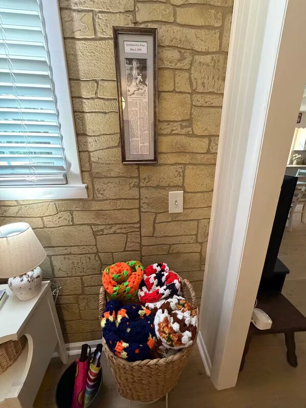 A room with a stone wall, a framed historical plaque, a basket of colorful crocheted items, and a lamp on a table.
