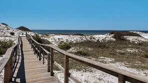 Ein hölzerner Steg führt zu einem Strand mit Dünen und blauem Himmel.