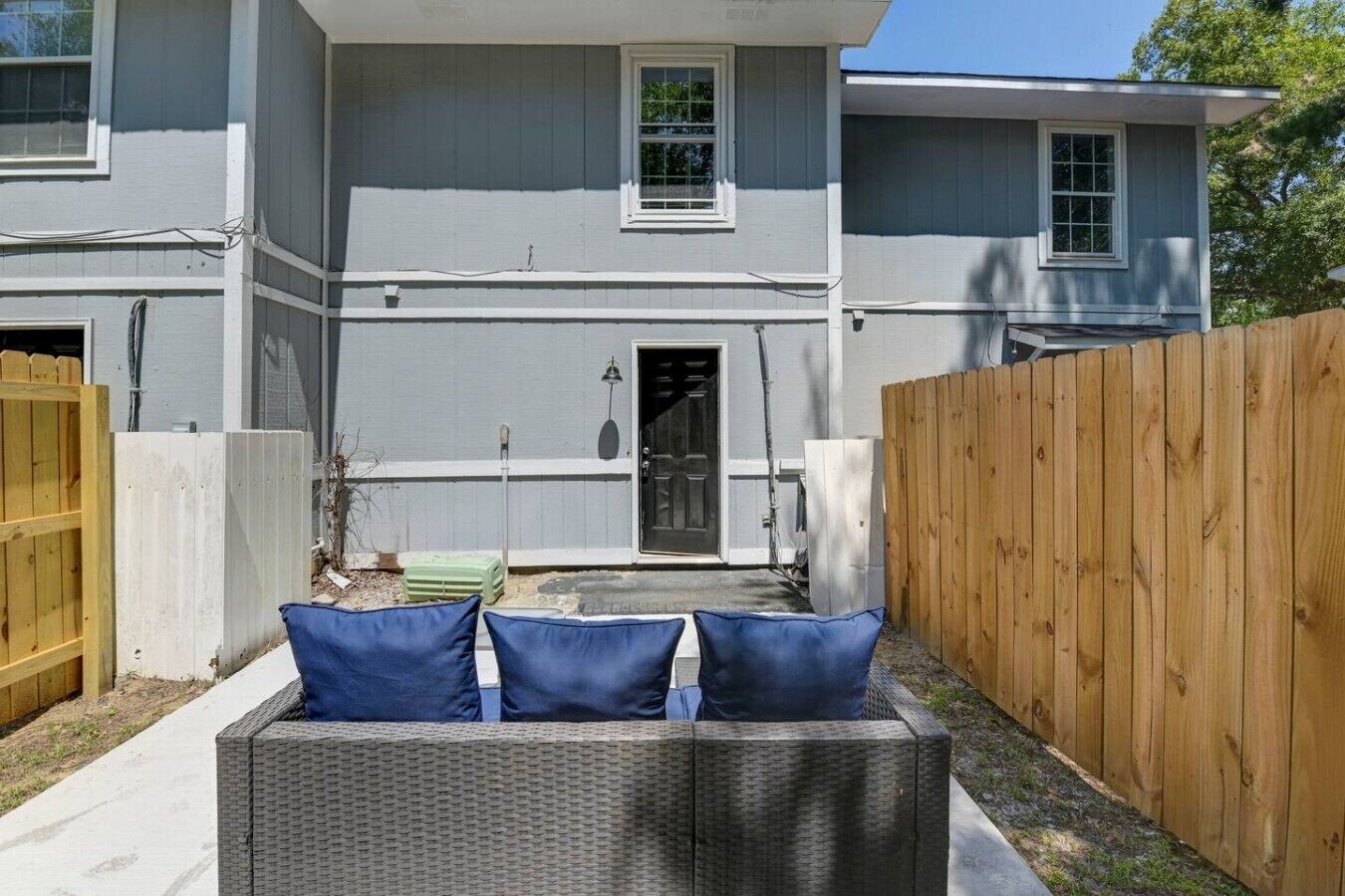 Una propiedad residencial con fachada gris, cerca de madera y un patio con sofá de mimbre y cojines.