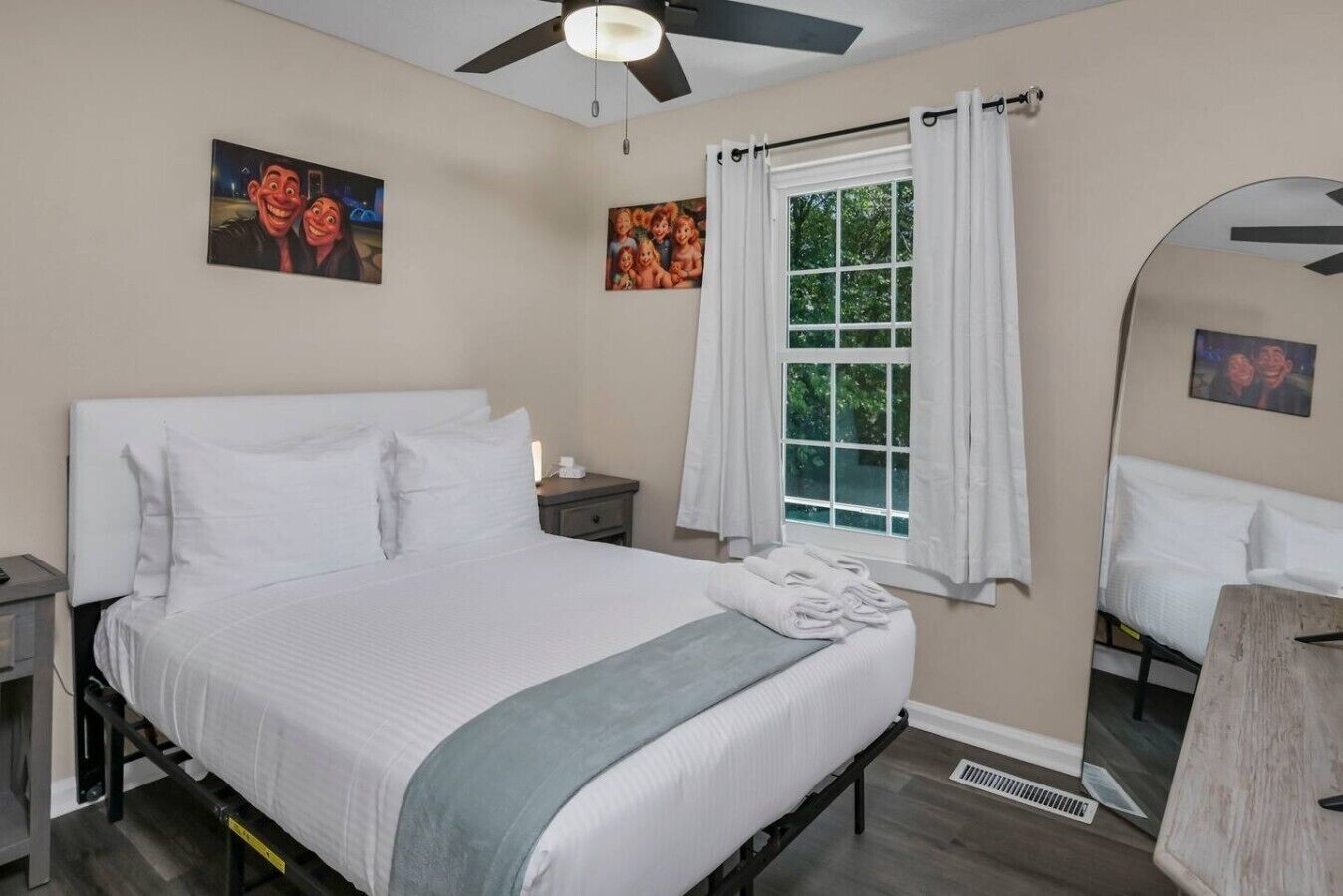 Ein Hotelzimmer mit einem Bett, einem Nachttisch, einem Fenster mit Vorhängen, einem Deckenventilator und Kunstwerken an den Wänden.