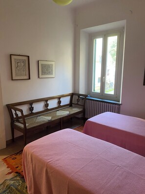 A room with a wooden bench, two beds with pink bedding, a window with a view, and two framed pictures on the wall. - Porzione di villa con giardino privato e A/C (Senigallia)