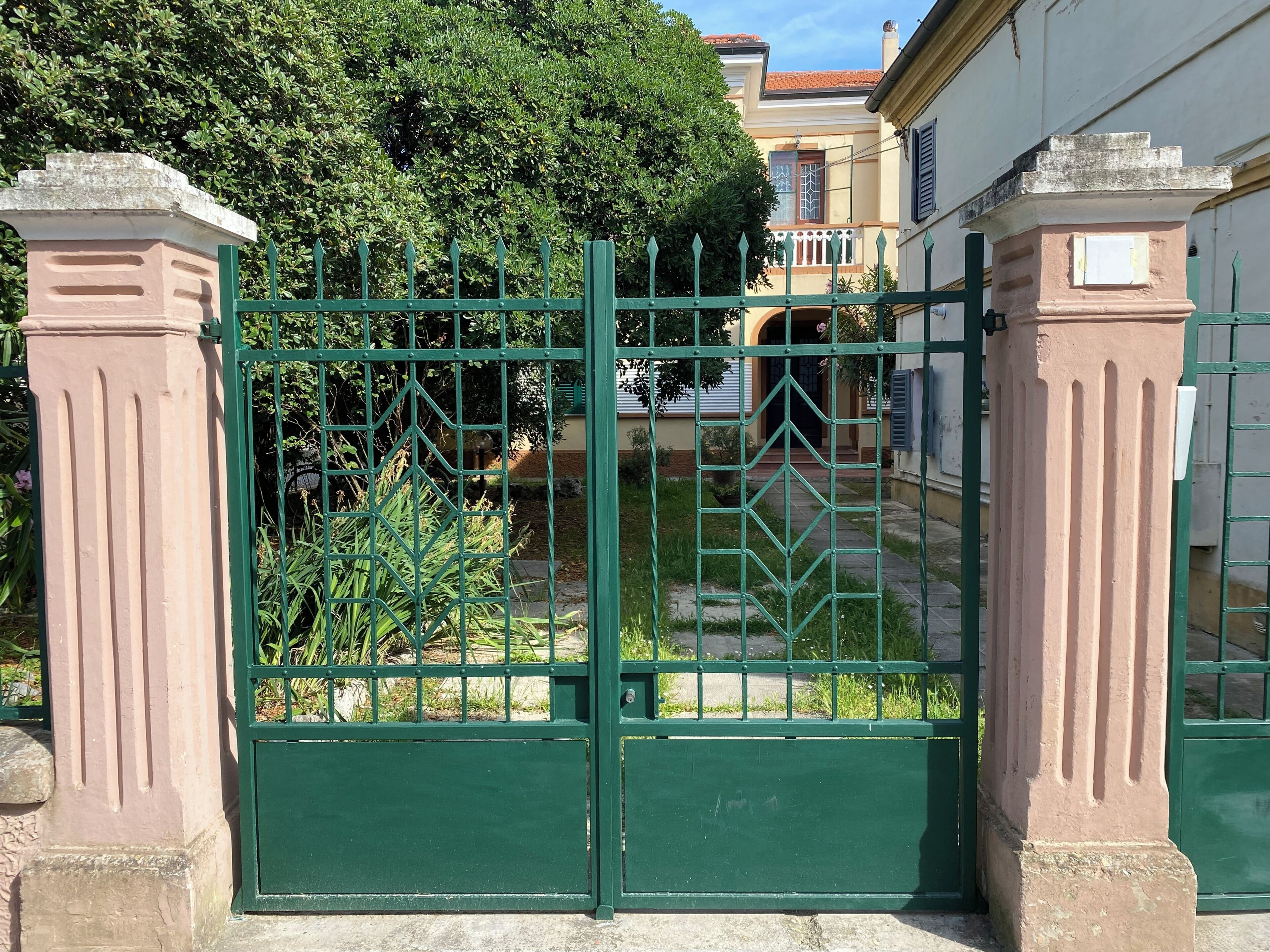 A gated entrance with a green metal gate, a stone pillar, and a building with a balcony in the background.