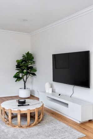 Una sala de estar moderna con una mesa redonda de madera, una planta en maceta, un mueble blanco para entretenimiento y un televisor de pantalla plana grande.