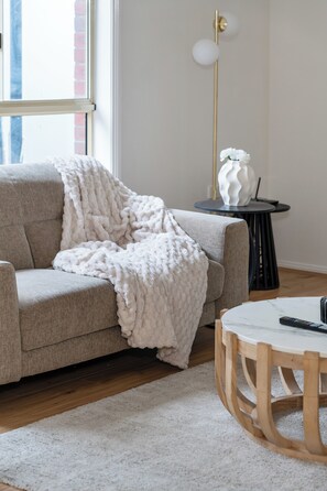 Una sala de estar con un sofá gris, una manta blanca, una mesa de centro redonda de madera y una mesita auxiliar negra con un jarrón blanco.