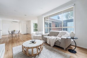 Ein großzügiger Wohnraum mit großem Fenster, einem runden Holztisch, einem grauen Sofa und einem Essbereich mit weißem Tisch und Stühlen.