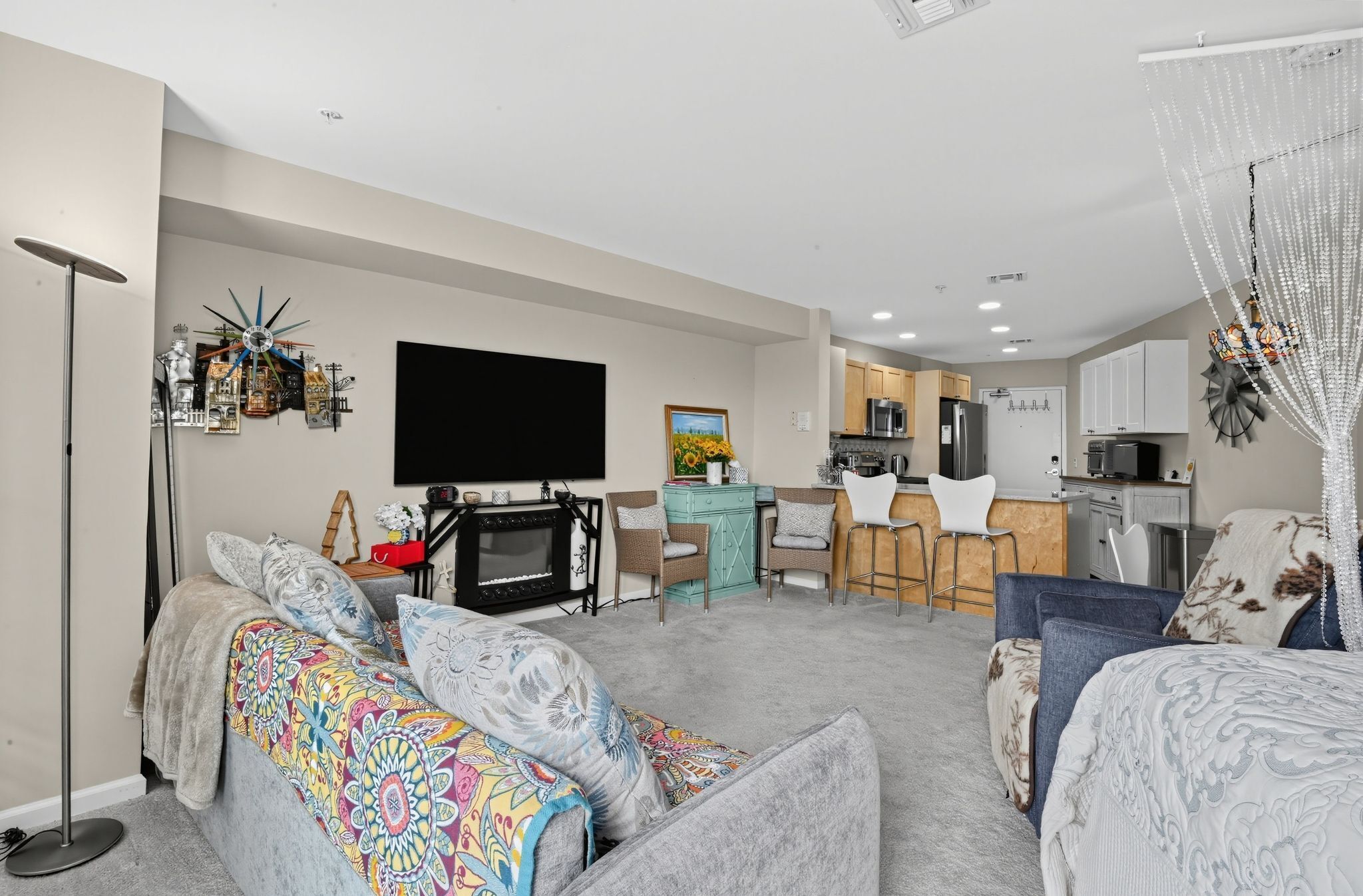 A modern living room with a flat-screen TV, a dining area with a kitchen in the background, and a cozy seating area with patterned cushions.