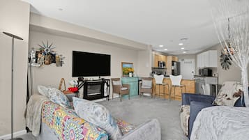 A modern living room with a flat-screen TV, a dining area with a kitchen in the background, and a cozy seating area with patterned cushions.