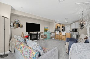 A modern living room with a flat-screen TV, a dining area with a kitchen in the background, and a cozy seating area with patterned cushions.
