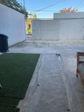 Eine Beton-Terrasse mit Kunstgrastürn, einer Metallmülltonne und einem Korbstuhl.