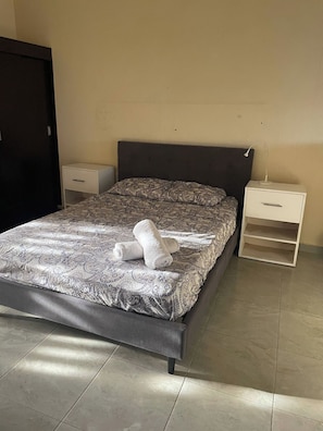 Ein Bett mit gemustertem Überwurf, ein Nachttisch mit Schublade sowie ein Beistelltisch mit Lampe.
