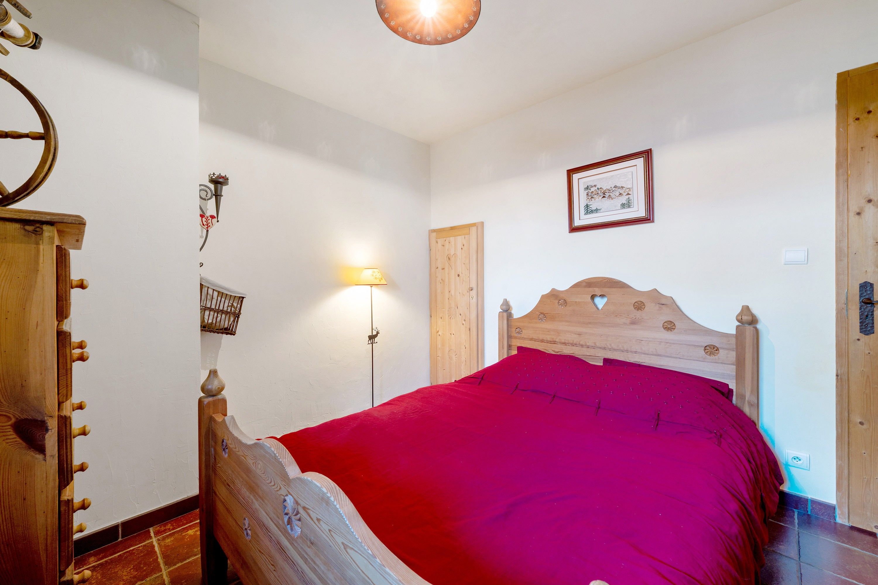 A bedroom with a wooden bed, a red bedspread, a wall-mounted lamp, and a framed picture.