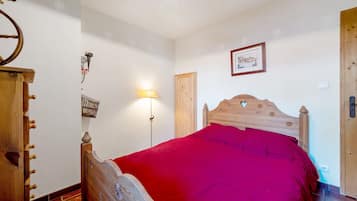 A bedroom with a wooden bed, a red bedspread, a wall-mounted lamp, and a framed picture.
