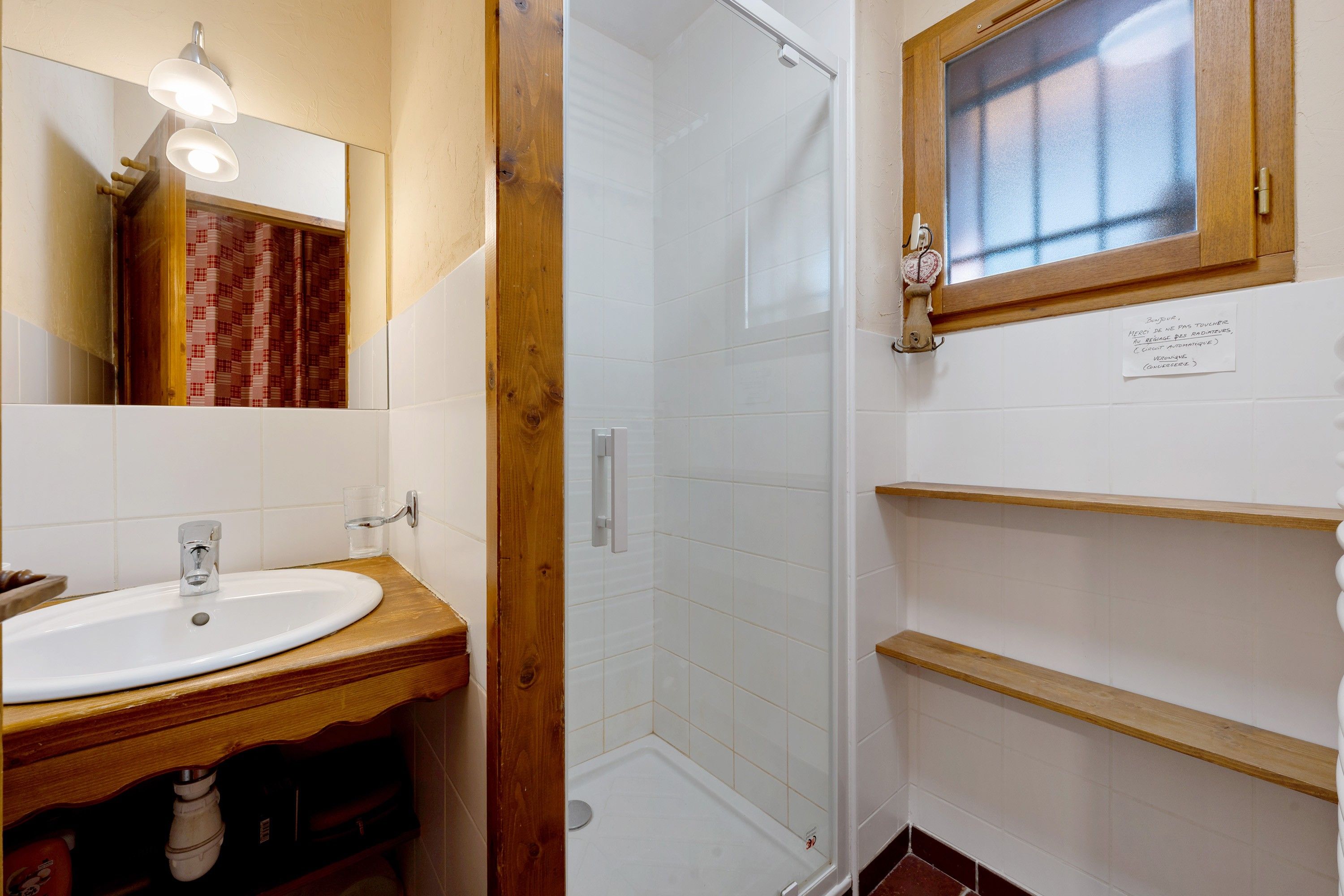 A bathroom with a wooden vanity, a mirror, a shower enclosure, and a small window.