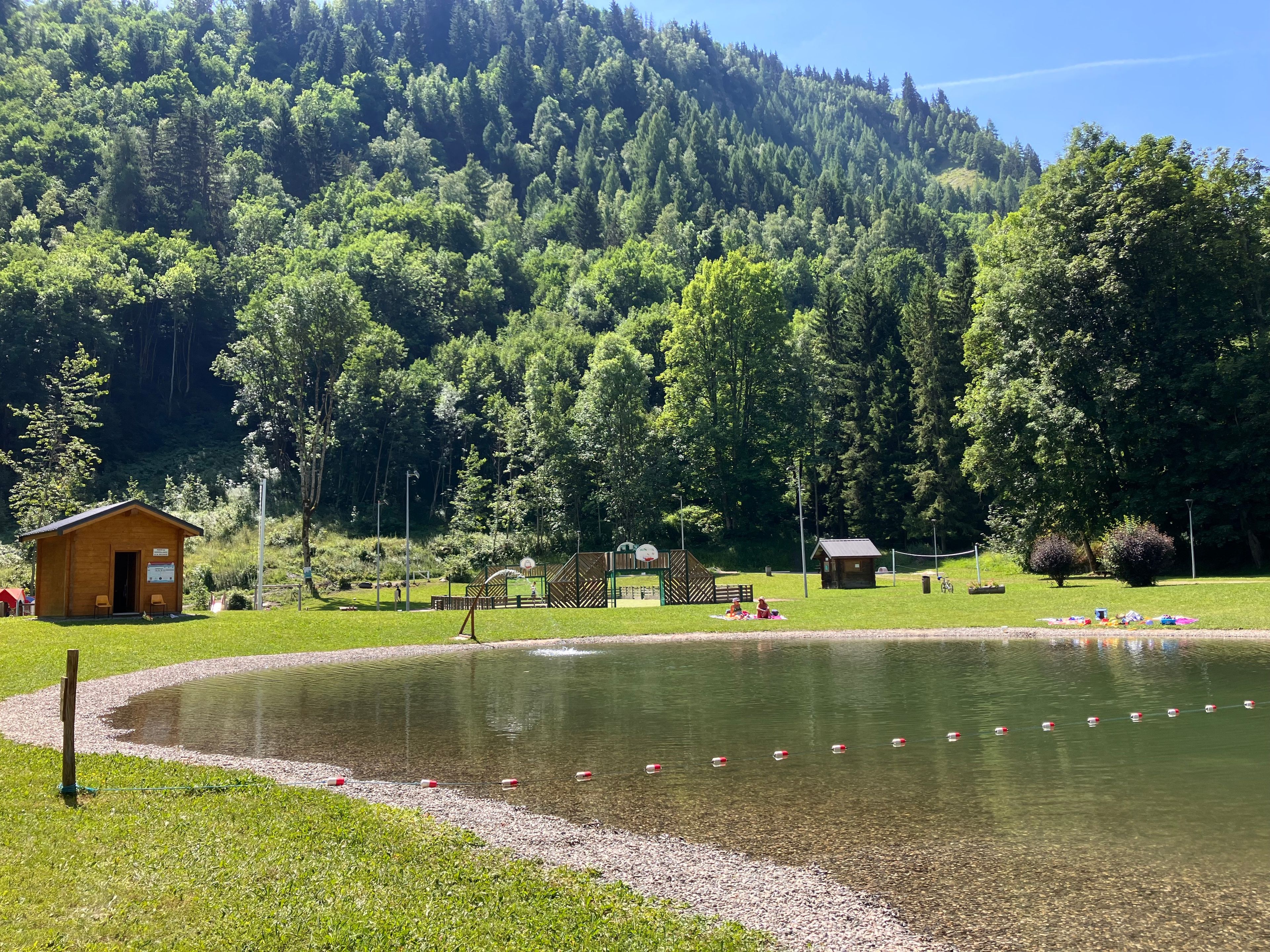 A lakeside park with a small wooden cabin, picnic tables, and a basketball court.
