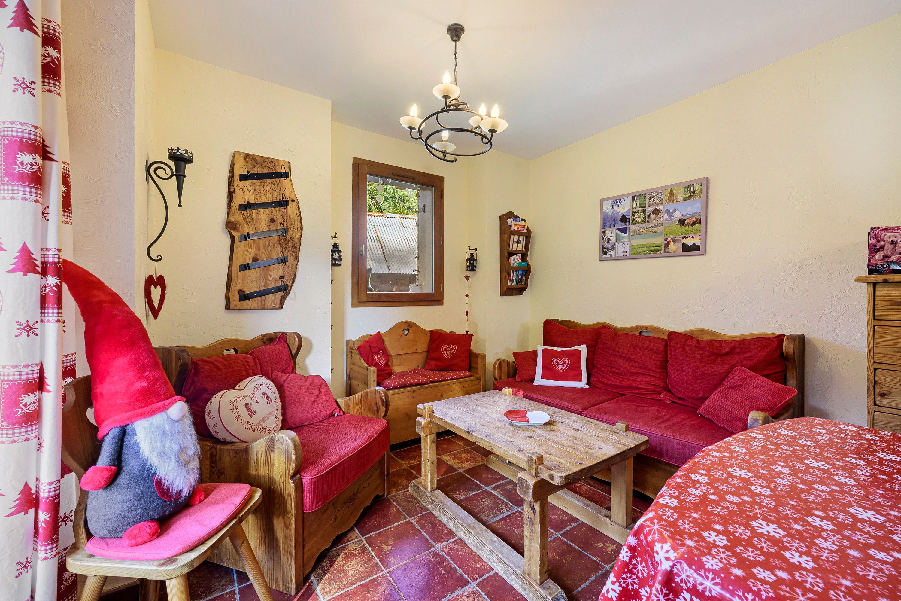 A cozy room with a wooden sofa, a wooden table, and a red chair. There is a window with a view of the outside, a chandelier, and a wall with a picture.