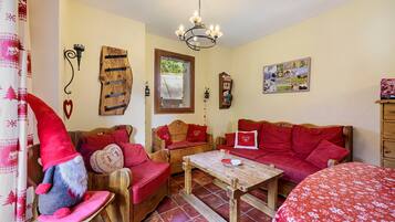 A cozy room with a wooden sofa, a wooden table, and a red chair. There is a window with a view of the outside, a chandelier, and a wall with a picture.