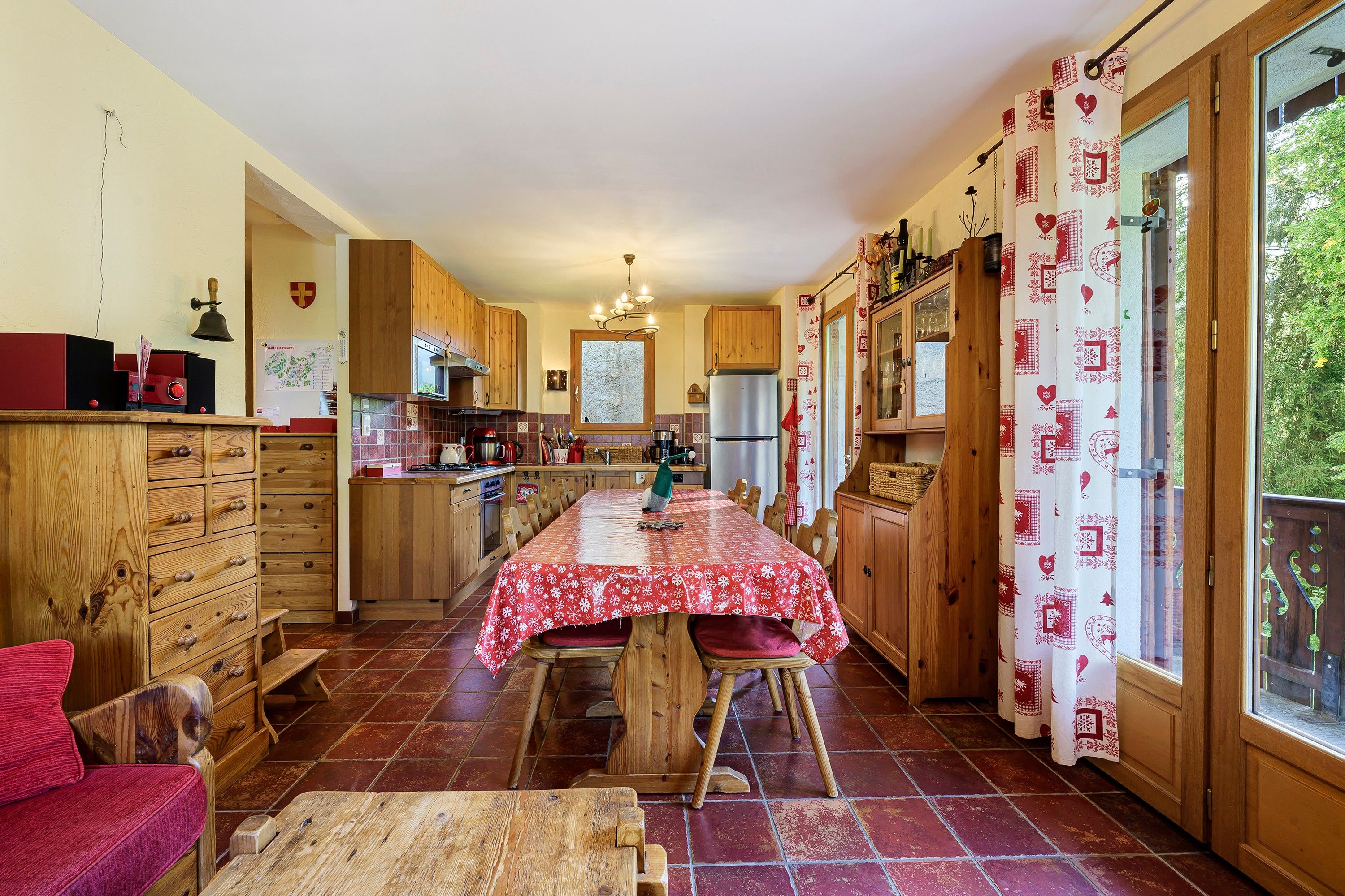 A kitchen with wooden cabinets, a dining table with a red tablecloth, and a view of greenery outside.