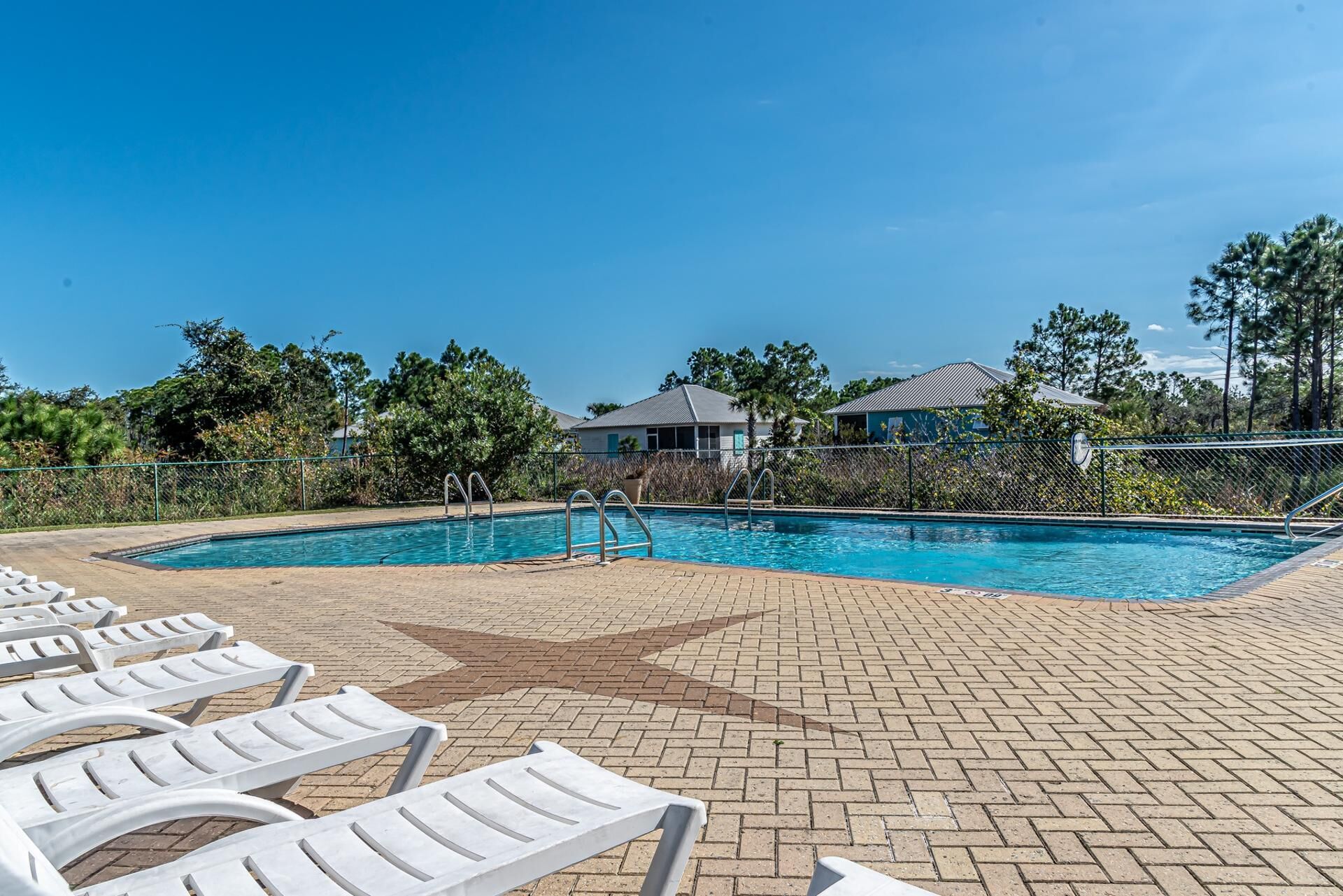 A swimming pool with lounge chairs, a brick patio, and a fenced area with trees and buildings in the background.