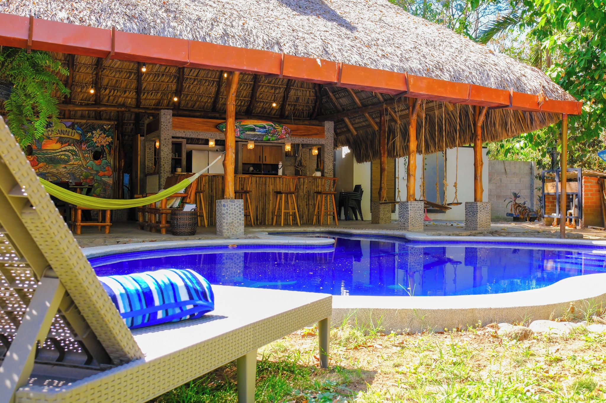 A pool area with a thatched roof structure, lounge chairs, and a bar area.