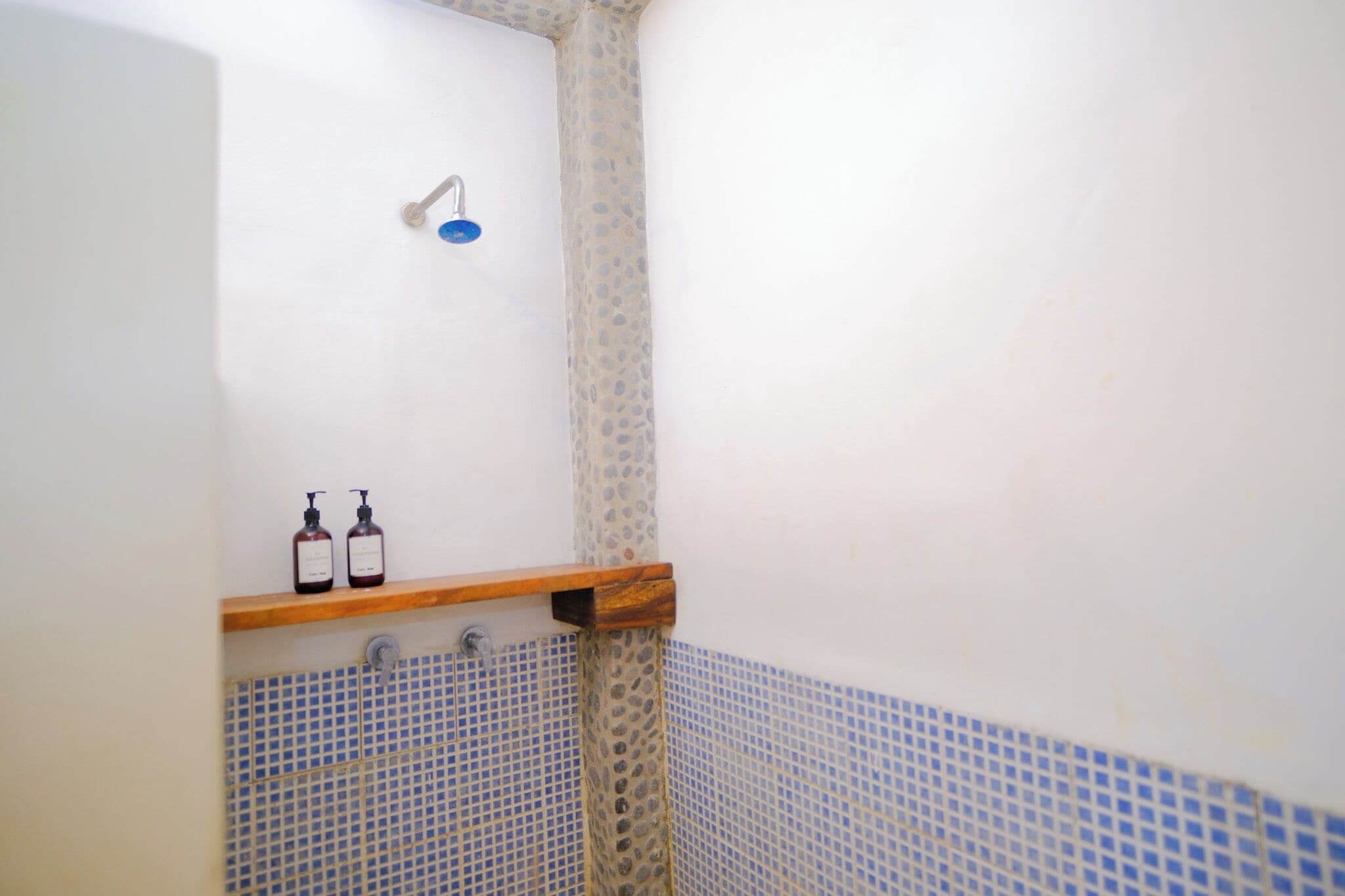 A bathroom with a shower, a wooden shelf with two bottles, and blue-tiled walls.