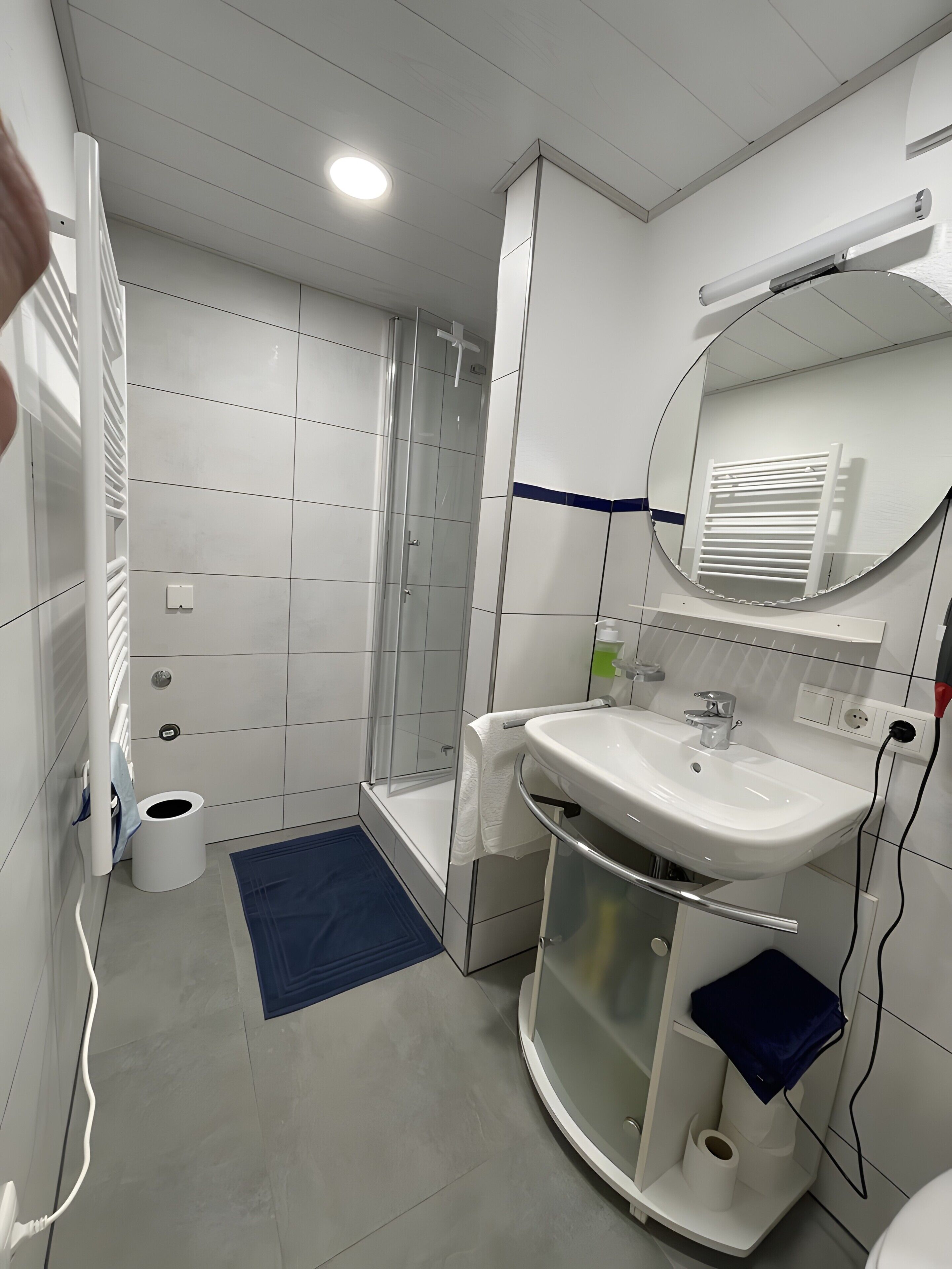 Ein modernes Badezimmer mit einer gläsernen Duschkabine, einem weißen Waschbecken mit Spiegel darüber und einer blauen Fußmatte auf dem Boden.