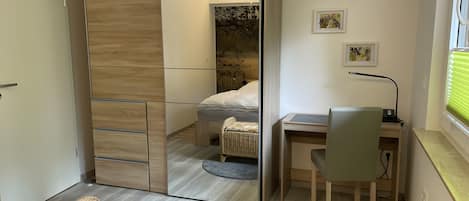 A modern bedroom with a mirrored wardrobe, a bed with white bedding, a desk with a chair, and artwork on the wall.
