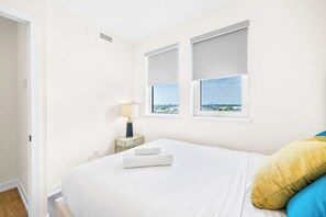 A hotel room with a bed, two windows, a nightstand, and a lamp. - Stylish 3BR+Den Apartment w/ Balcony, Views & Free Parking (Toronto)