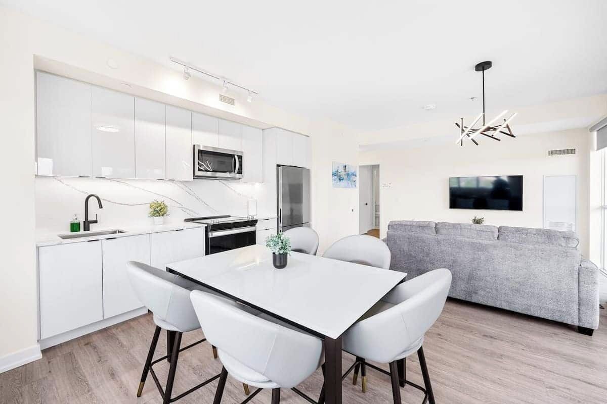 A modern kitchen with white cabinets, a dining table with four chairs, and a grey sofa in the living area.
