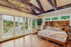 A bedroom with a large bed, wooden beams, and a view of greenery through large windows. - 3BR waterfront home with dock and hot tub (Bainbridge Island)