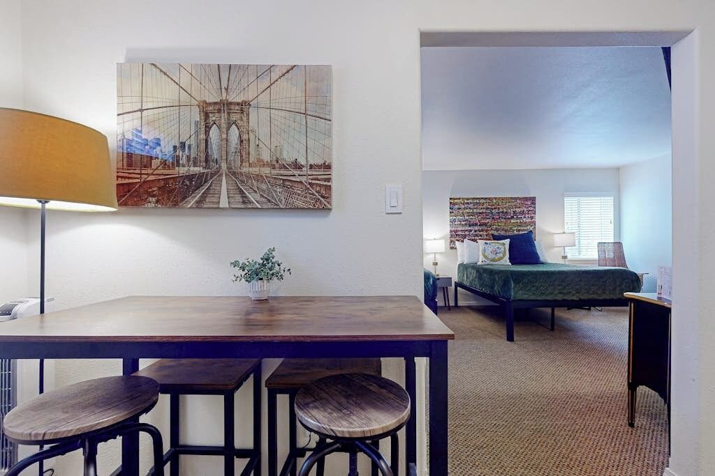 A room with a bed, a desk with stools, a lamp, and a painting of a bridge.