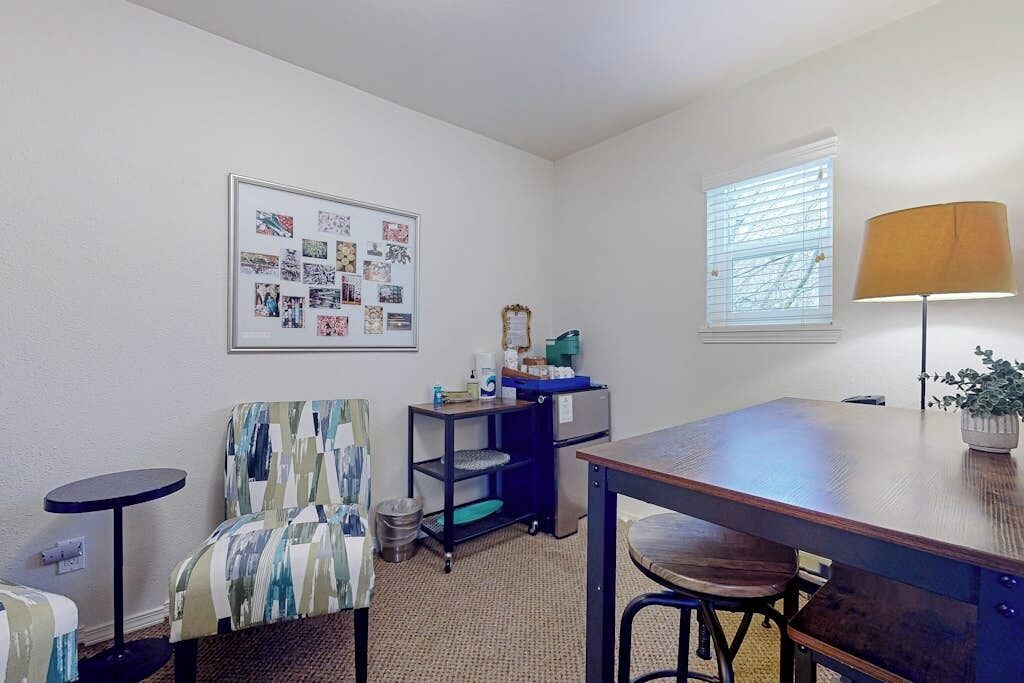 A room with a dining table, chairs, a small round table, a wall-mounted shelf, a trash bin, a window with blinds, and a framed collage on the wall.