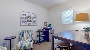A room with a dining table, chairs, a small round table, a wall-mounted shelf, a trash bin, a window with blinds, and a framed collage on the wall.