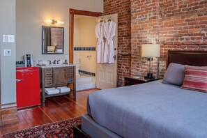 A hotel room with a bed, a red minibar, a wooden dresser, a bathroom with a shower, and a brick wall.