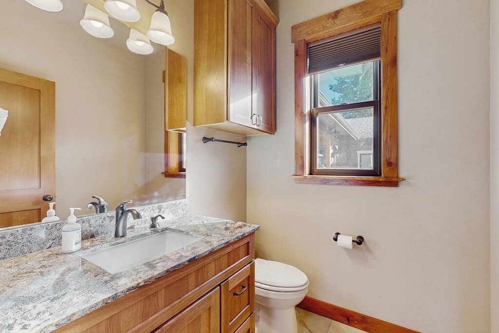 A bathroom with a marble sink, wooden cabinets, a toilet, and a window with blinds.