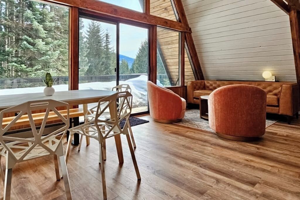 Ein gemütliches Interieur mit einem Holztisch, Stühlen, einer Couch und einem großen Fenster mit Blick auf eine verschneite Landschaft.