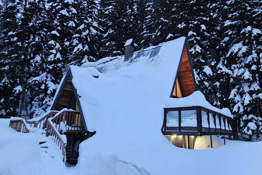 Une cabane recouverte de neige, avec un toit à forte pente, un revêtement extérieur en bois et un escalier menant à une terrasse.