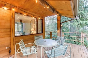 A wooden deck with a round table and chairs, a window with a view of greenery, and a hanging light fixture. - Stylish 3BR - Fireplace - Deck - Washer Dryer (Port Angeles)