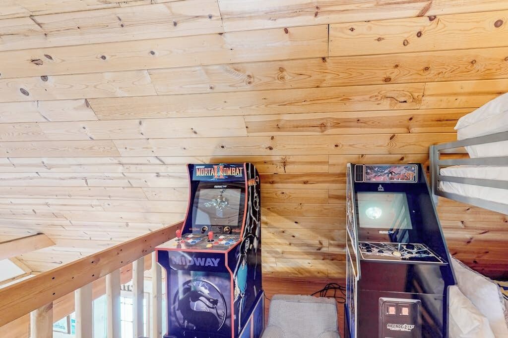 A room with wooden ceiling and walls, featuring two arcade machines, a bunk bed, and a window.