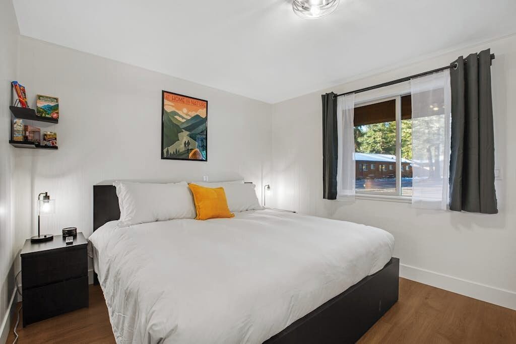A bedroom with a bed, a nightstand, a window with curtains, and a wall-mounted shelf with books.