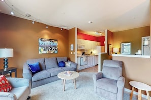 A modern living room with a grey sofa, armchair, and coffee table. A kitchen with white cabinets and red accents is visible in the background.