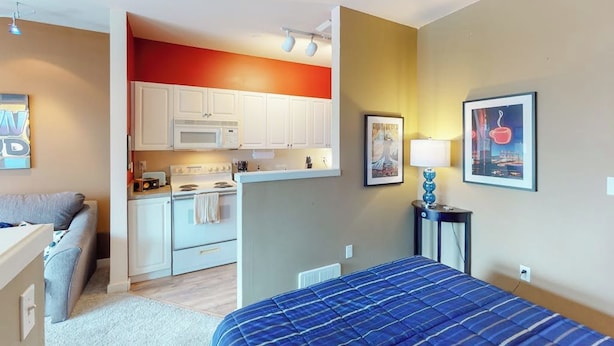 Een compact hotelkamer met kitchenette, een bed met blauwe deken, een klein zithoekje en een muur met twee ingelijste foto's.