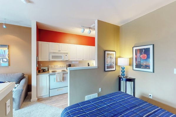 Une chambre d’hôtel compacte comprenant un coin cuisine, un lit avec une couverture bleue, un petit coin salon et un mur orné de deux tableaux encadrés.