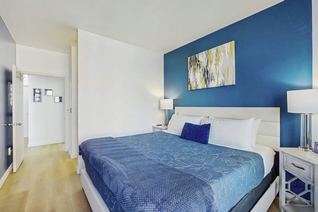 A bedroom with a bed, a nightstand, a wall-mounted painting, and a door leading to another room.