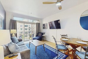 A modern living room with a sofa, coffee table, dining table, and chairs. There is a TV mounted on the wall, a ceiling fan, and a large window with curtains. - Dog-Friendly Condo with Heated Pool, Hot Tub, Balcon (Seattle)