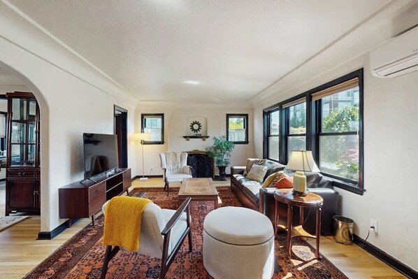 A spacious living room with a sofa, armchair, and a television. There are large windows providing natural light and a view outside. - 2 BR Home in the heart of Beacon Hill (Seattle)
